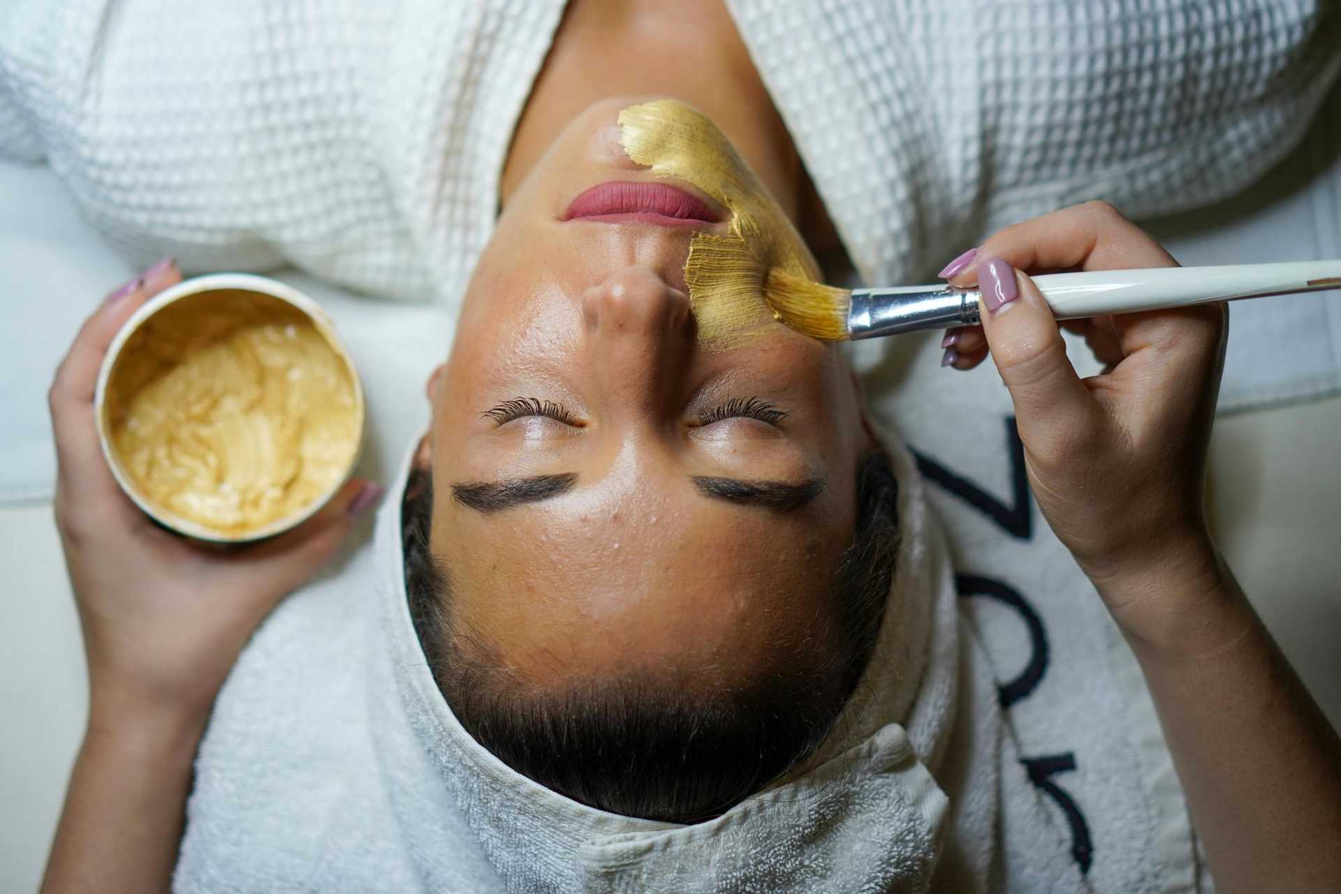 Woman receiving a facial treatment with a brush applied mask, eyes closed, wearing a white robe.