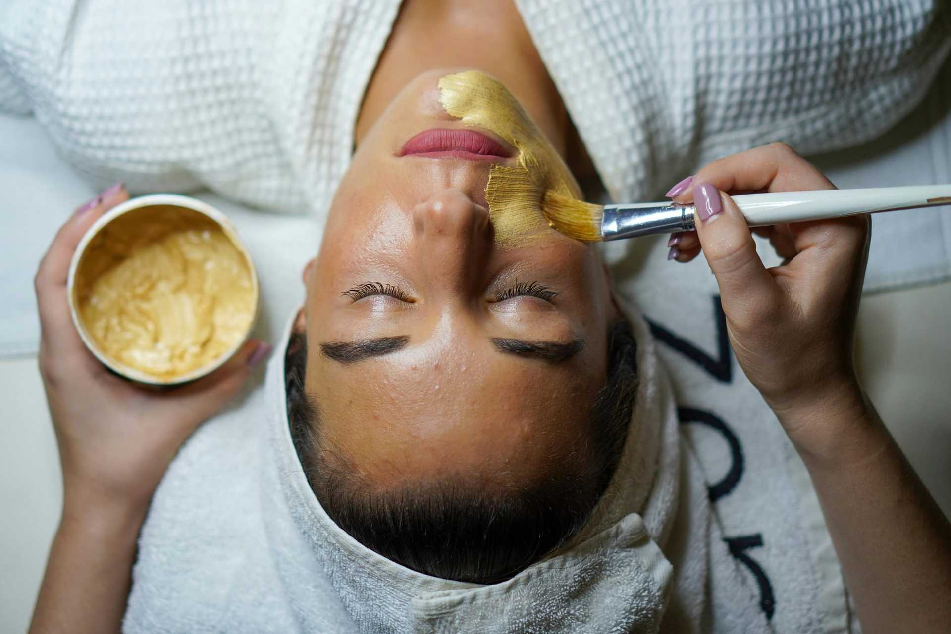 Woman receiving a facial treatment with a brush applied mask, eyes closed, wearing a white robe.