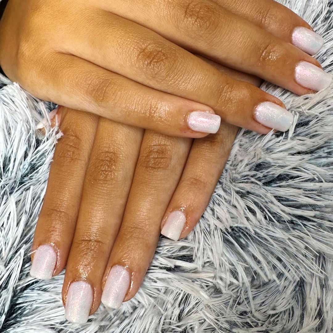 Close-up of hands with iridescent, shimmery white nail polish resting on a fluffy gray surface.