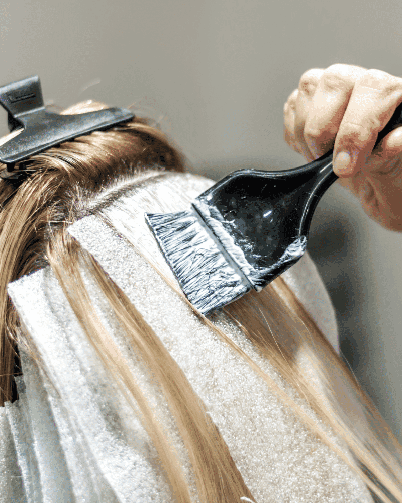 Applying hair dye with a brush on blonde hair, using foils to separate sections for highlights.